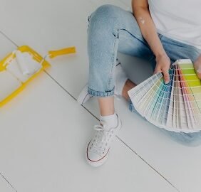 Faceless woman in jeans holds color samples, chooes best tone for refurbishment walls, sits on white floor, uses tray and paint roller for house remodeling. Home makeover and renovation concept
